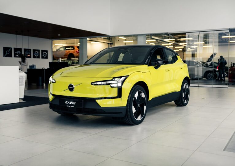 Ein moosgelber Volvo EX30 steht in einem modernen Autohaus mit großen Glasflächen und heller Beleuchtung. Das Elektro-SUV ist aus der Frontperspektive fotografiert, mit schwarzen Designelementen an Dach und Frontpartie. Im Hintergrund sind weitere Fahrzeuge und Personen im Showroom zu sehen.